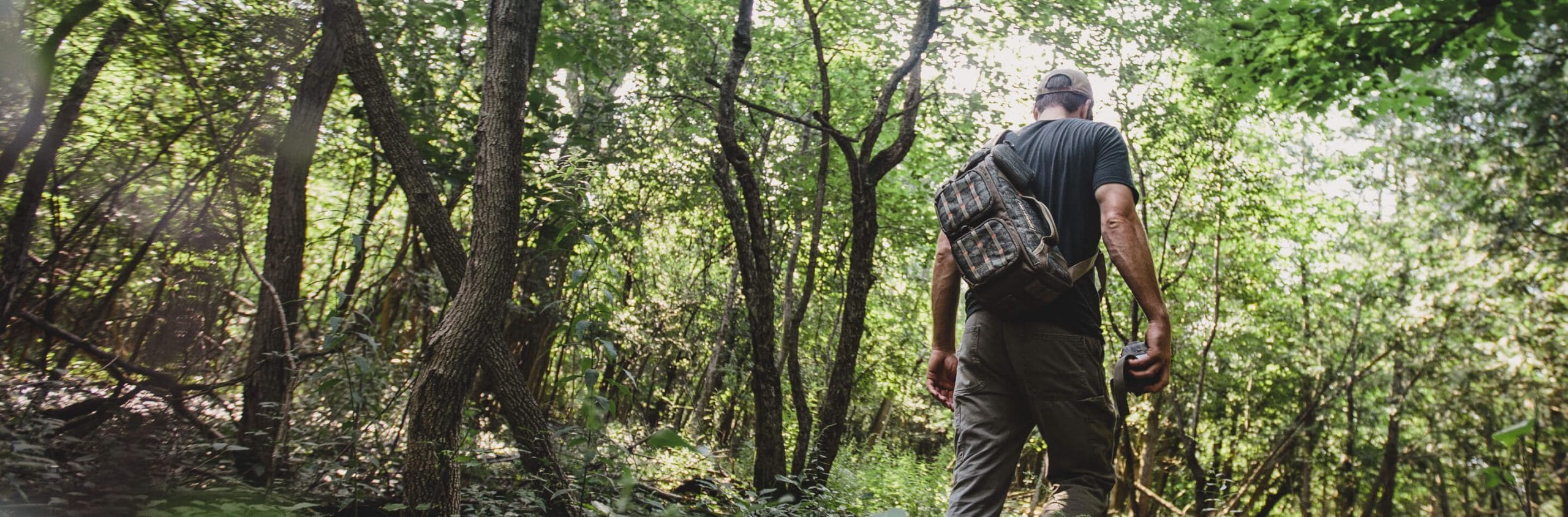 Man with camo trail camera carrier on his back walking through the woods