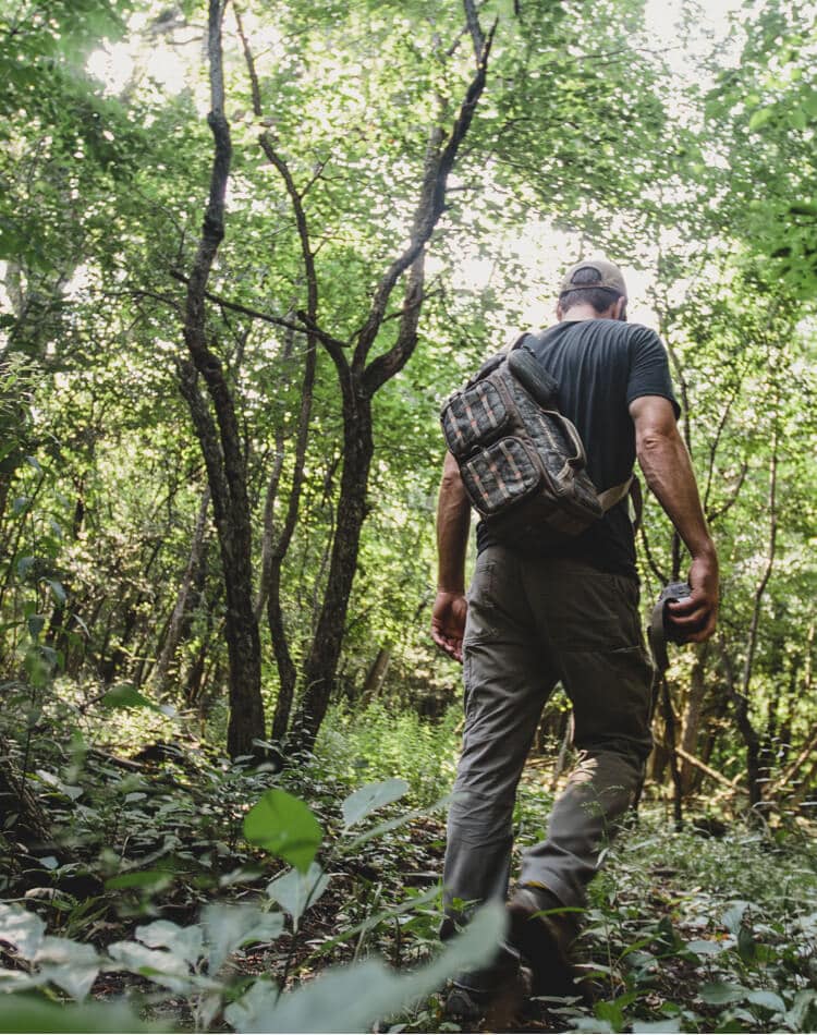 Man with camo trail camera carrier on his back walking through the woods