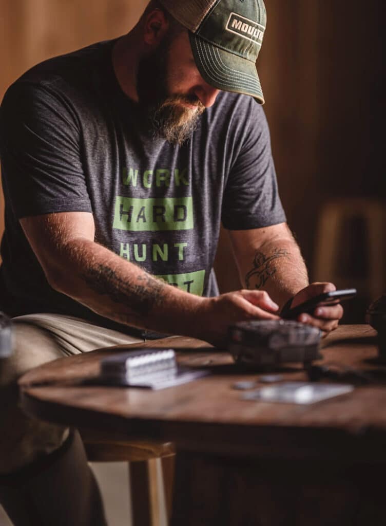 Man wearing Moultrie hat sitting at a table look at his phone next to his cellular trail camera